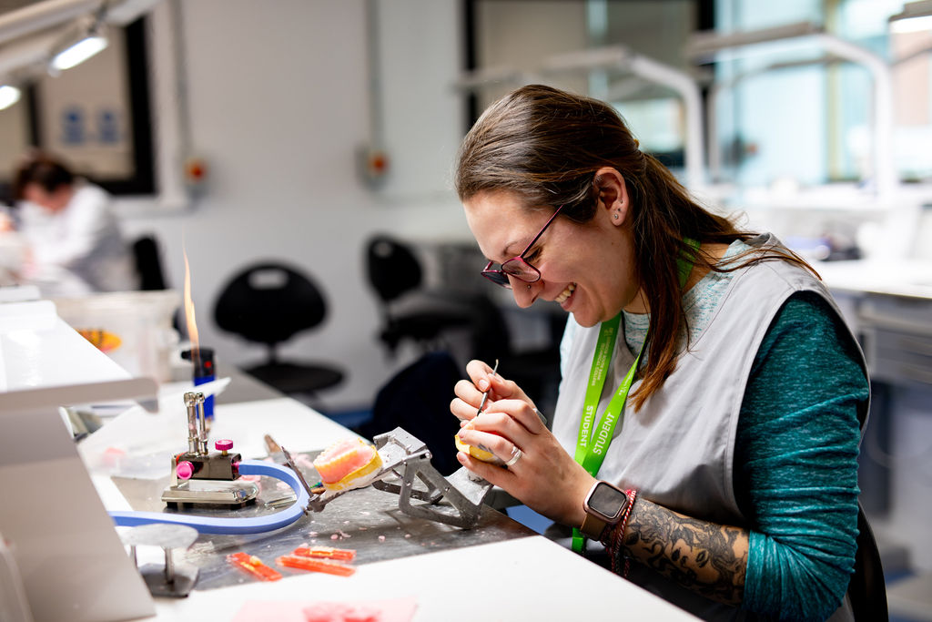 A LSBTC Dental student wearing glasses and a lanyard is carefully working with tools and small objects at a crafting bench, illuminated by a small flame and work lights in a well-organised, modern dental workshop.